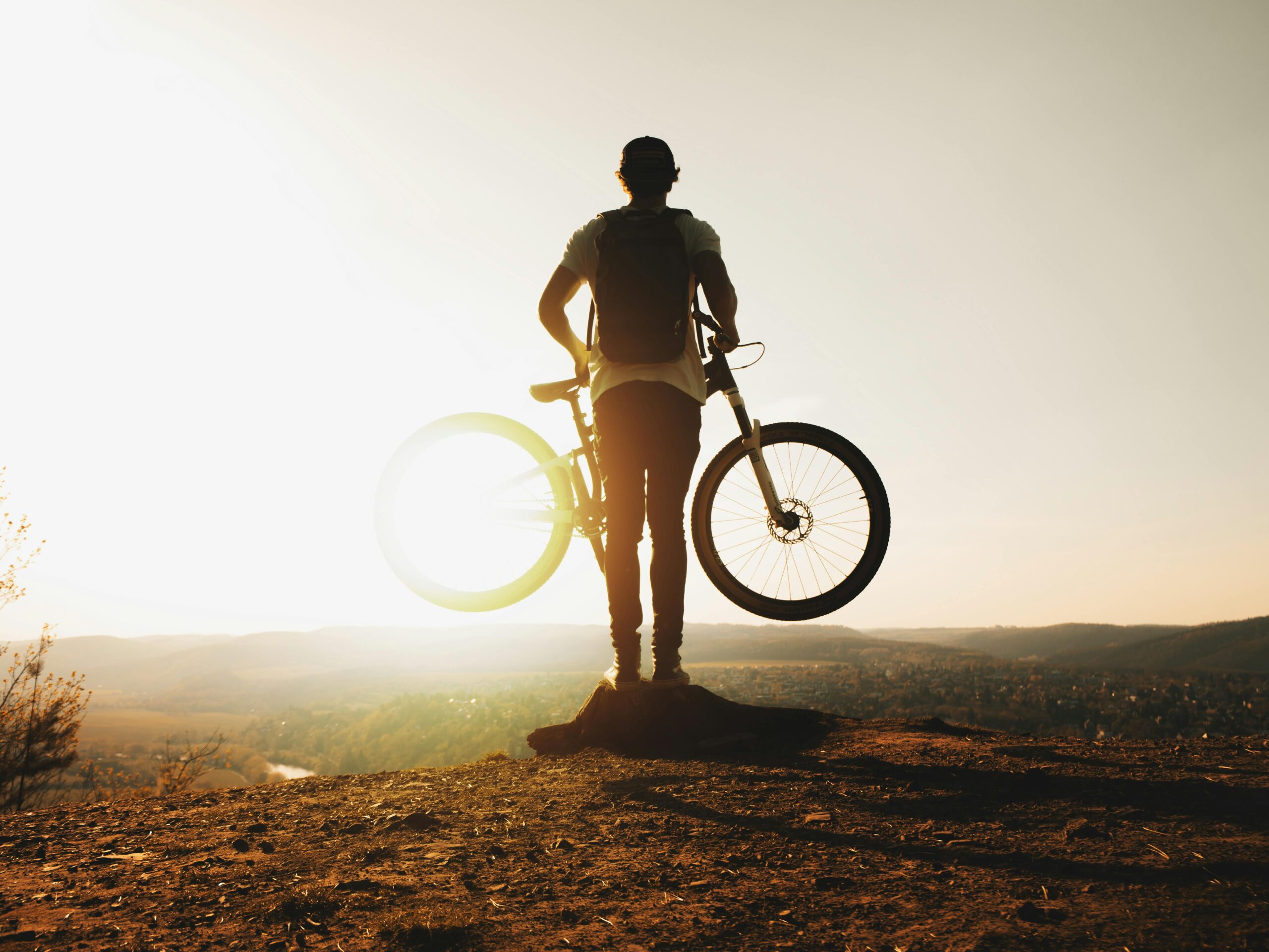 A cyclist stands silhouetted with a mountain bike on a hilltop during sunset, evoking adventure and freedom.